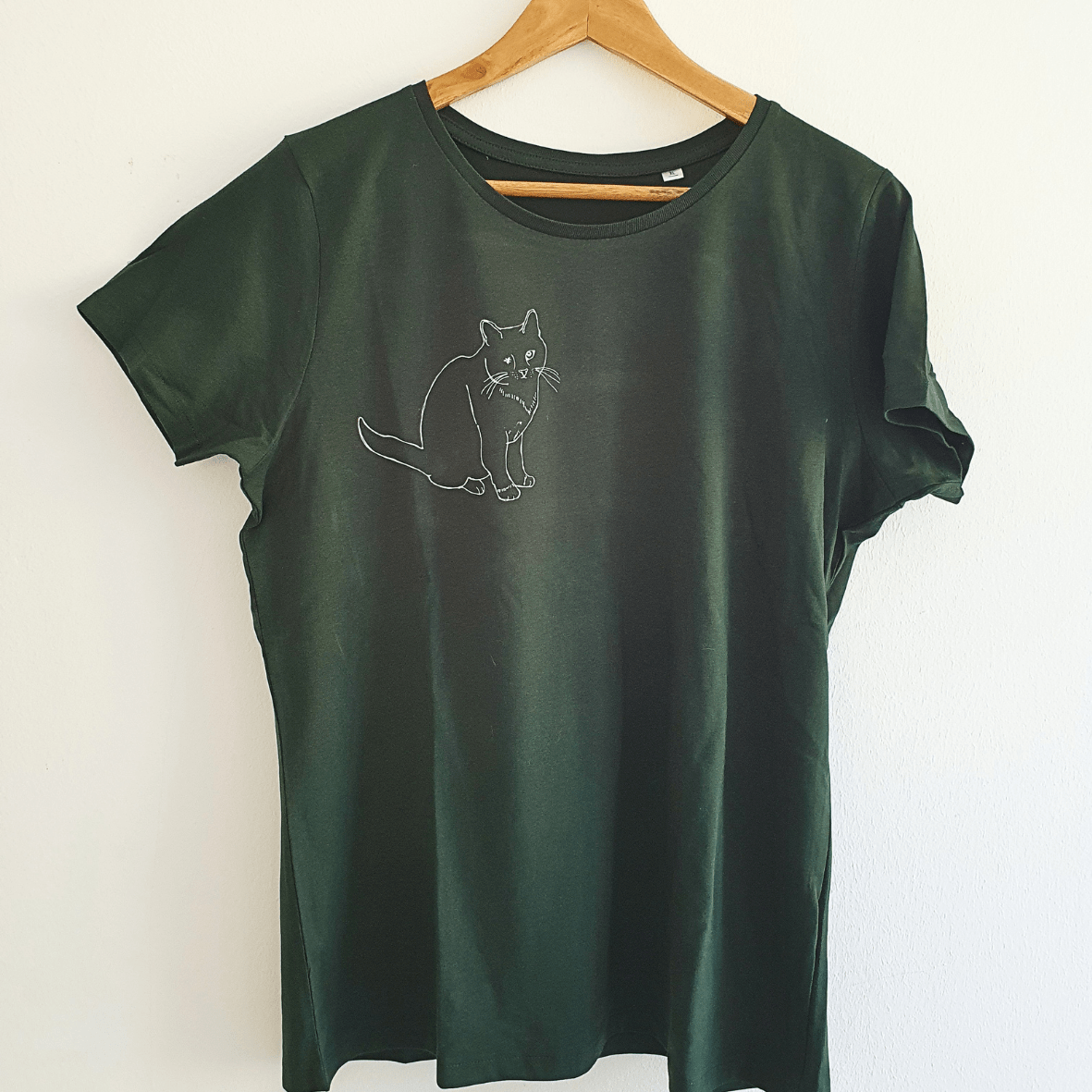 Ein grünes T-Shirt hängt an einem Kleiderbügel vor einer weißen Wand. Auf dem Shirt ist mit Siebdruck in Lineart in weiß eine einäugige Katze aufgedruckt.