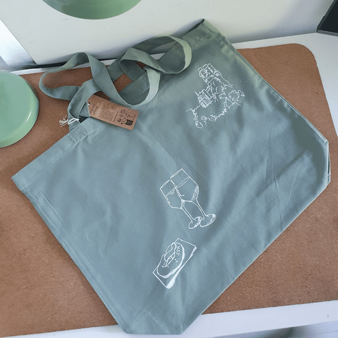 Organic Maxi Shopper Bag grün mit weißem Siebdruck, der in Lineart zwei Gläser Wein, eine Brezel und ein eine Burg am Rheinufer zeigt. Die Tasche liegt auf einer Korkmatte auf einem weißen Schreibtisch.